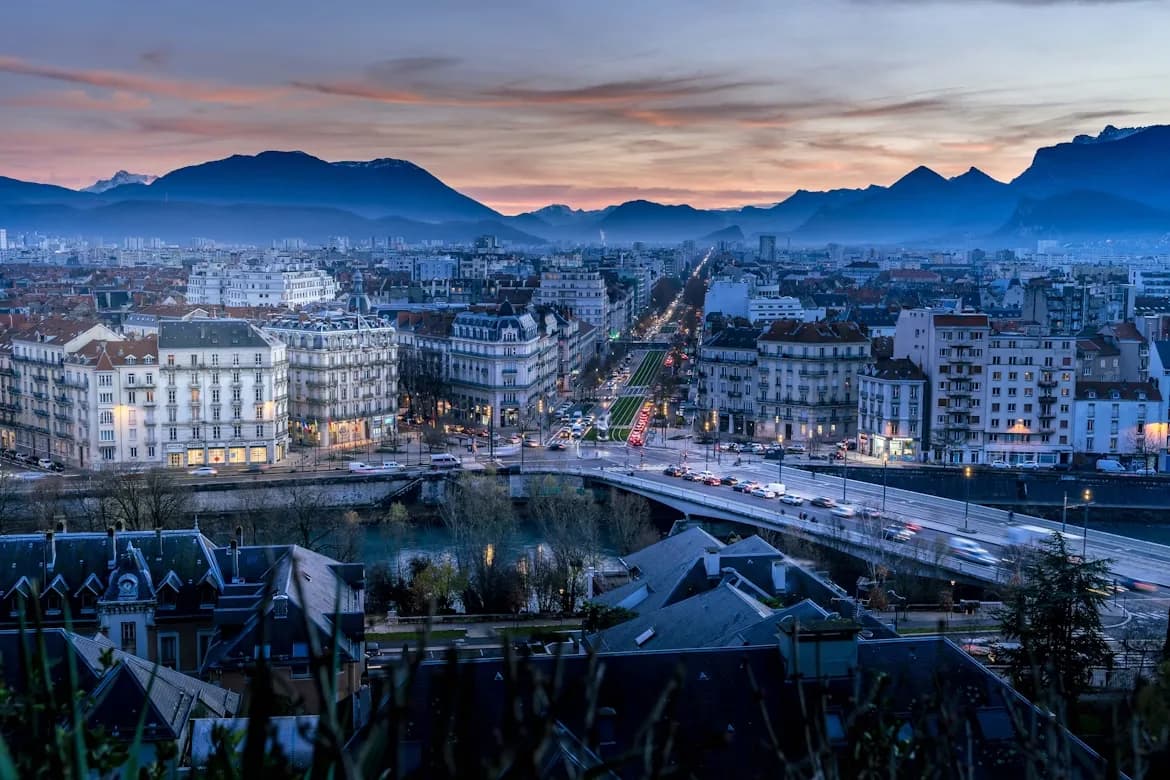 The city of Grenoble glows at dusk, with traffic lights creating streaks of light on a bridge as the mountains are silhouetted against the sky.