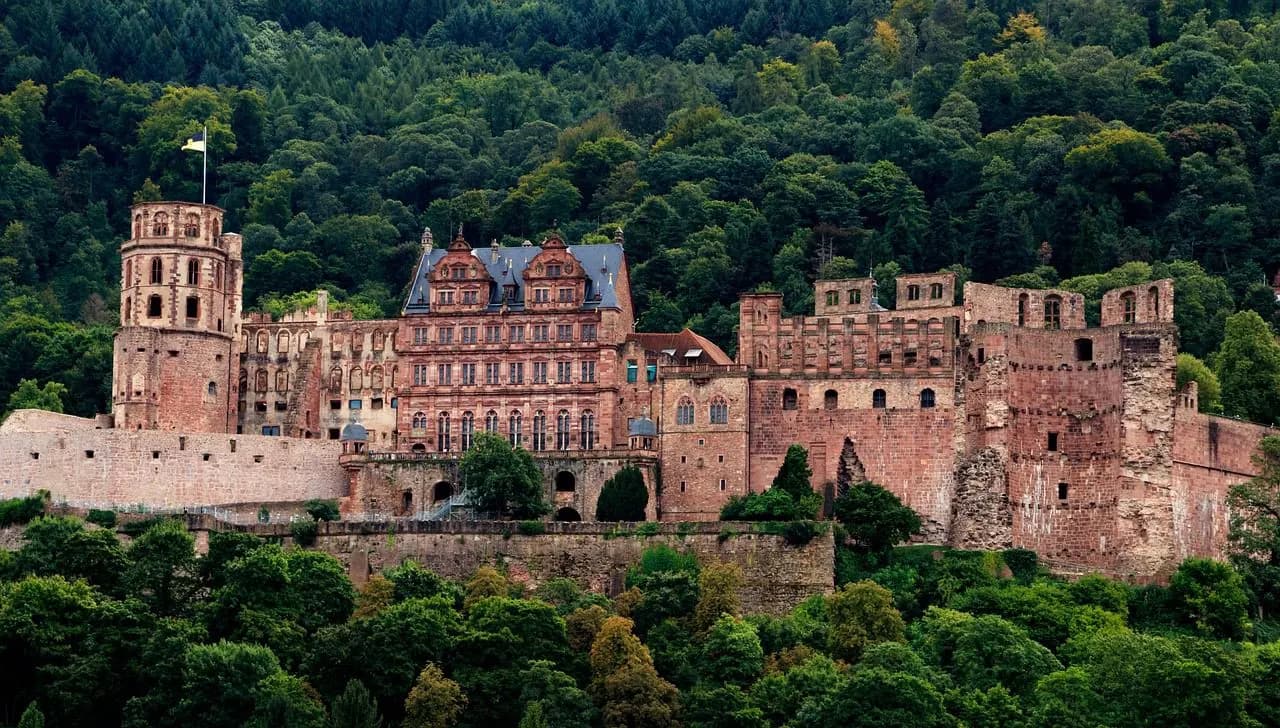 The dramatic red sandstone ruins of Heidelberg Castle are beautifully framed by lush green forests on the hillside.