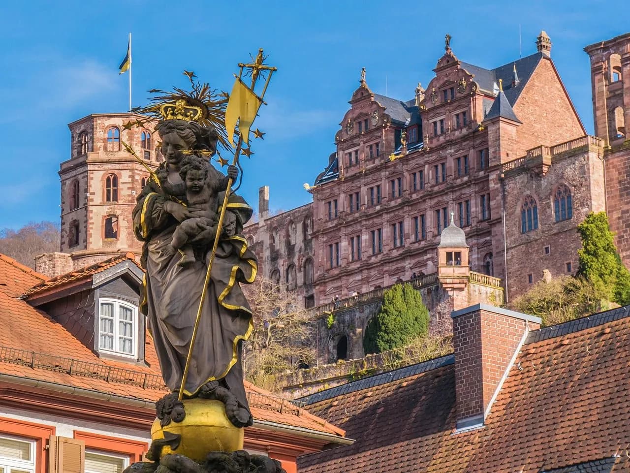 A historic fountain with a statue of the Virgin Mary stands in the foreground, with the famous Heidelberg Castle visible on the hill behind it.