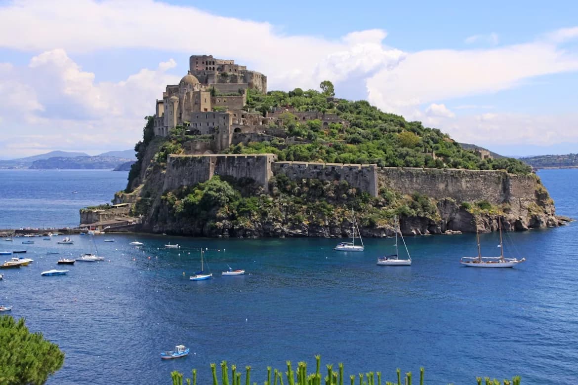The iconic Aragonese Castle rises from the sea, surrounded by boats and greenery under a blue sky.