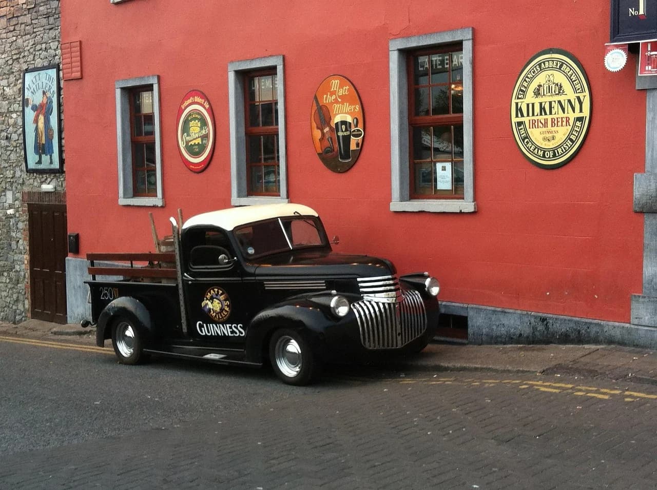 A vintage black truck with "Guinness" written on it is parked outside a red pub, with various signs and advertisements on the wall.