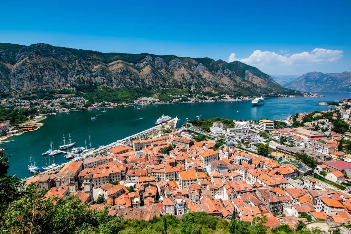 A high-angle view captures the medieval Old Town of Kotor, with its red rooftops and ancient walls nestled by the bay and dramatic mountains.