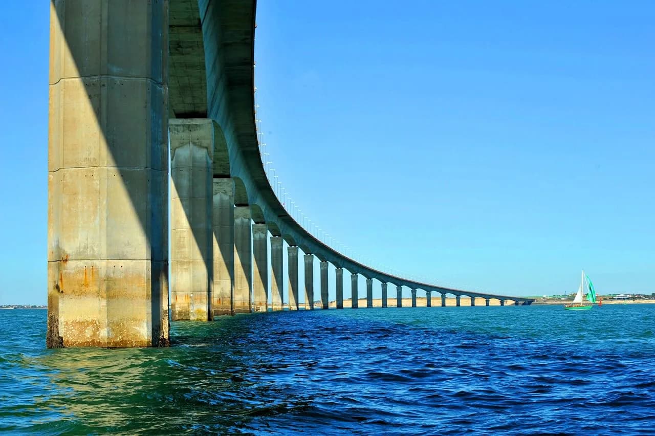 A sailboat glides under the impressive La Rochelle bridge, showcasing its soaring concrete arches against a clear blue sky.
