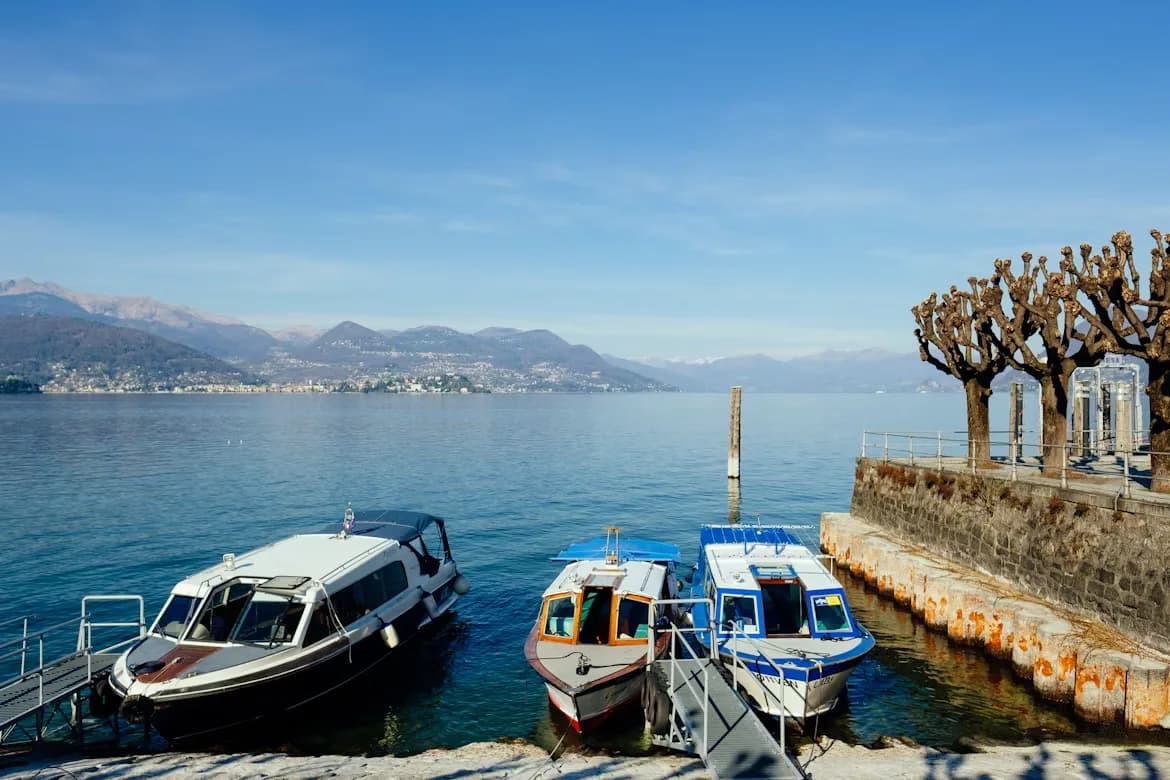 A selection of small boats is moored at the edge of Lake Orta, with a clear view of the surrounding mountains on a sunny day.