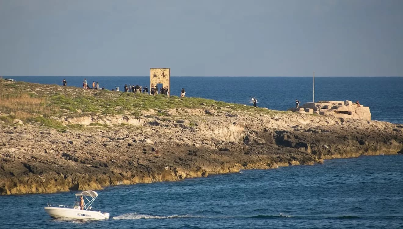 A group of people stands on a rocky cliff overlooking the sea, with a small monument and a boat in the distance.