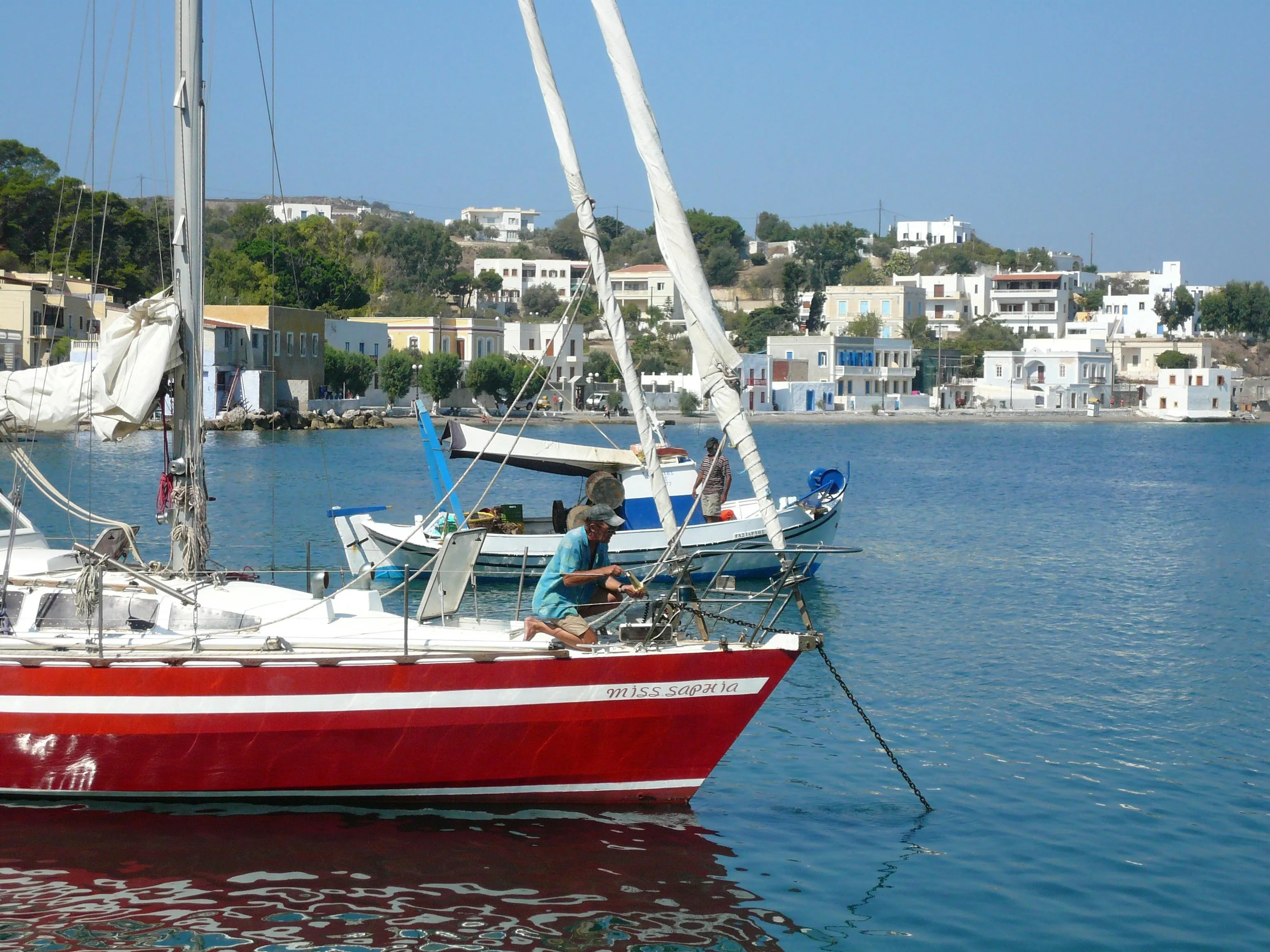 Boats are moored in a bay on the island of Leros, with a man working on a red sailboat in the foreground and traditional buildings on the shore.