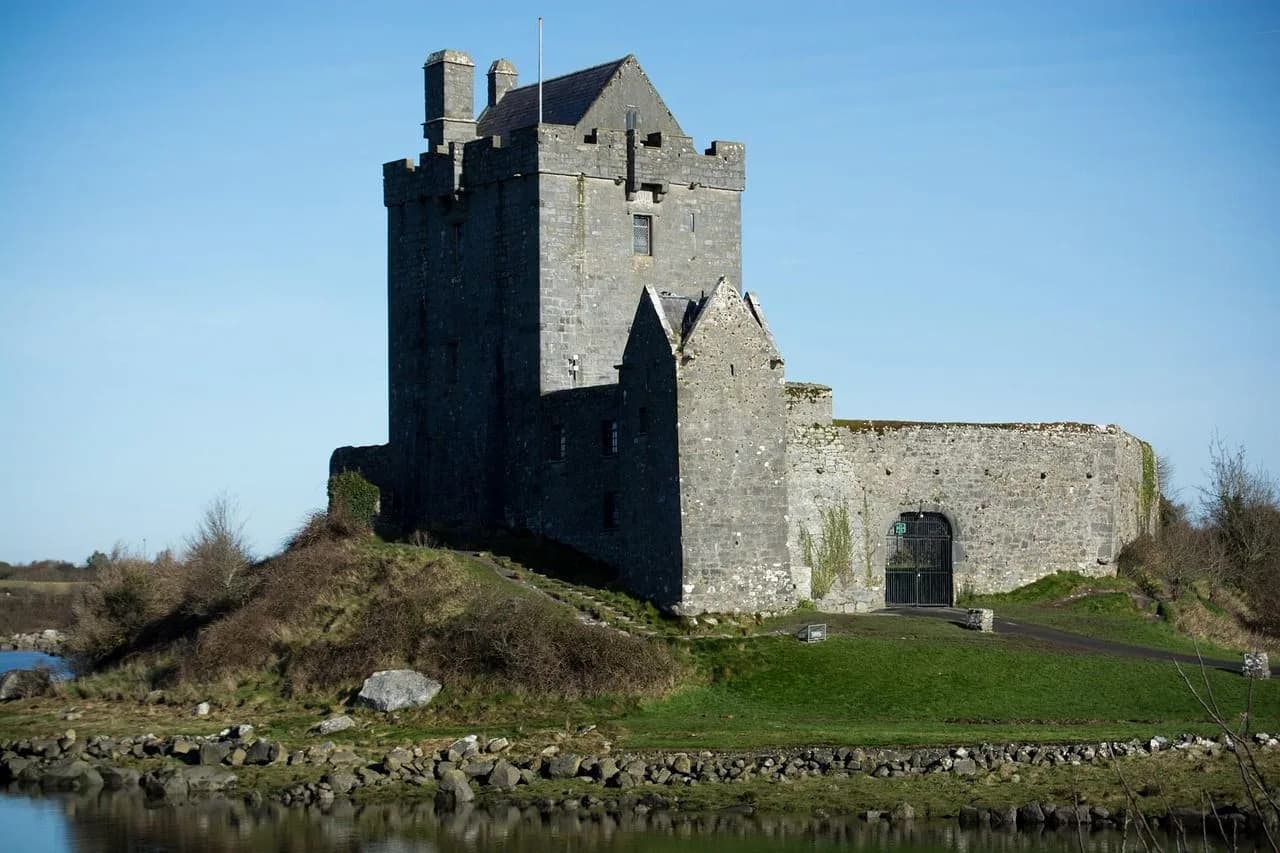 The medieval Bunratty Castle, a well-preserved fortress, stands on a grassy hill beside a tranquil body of water.