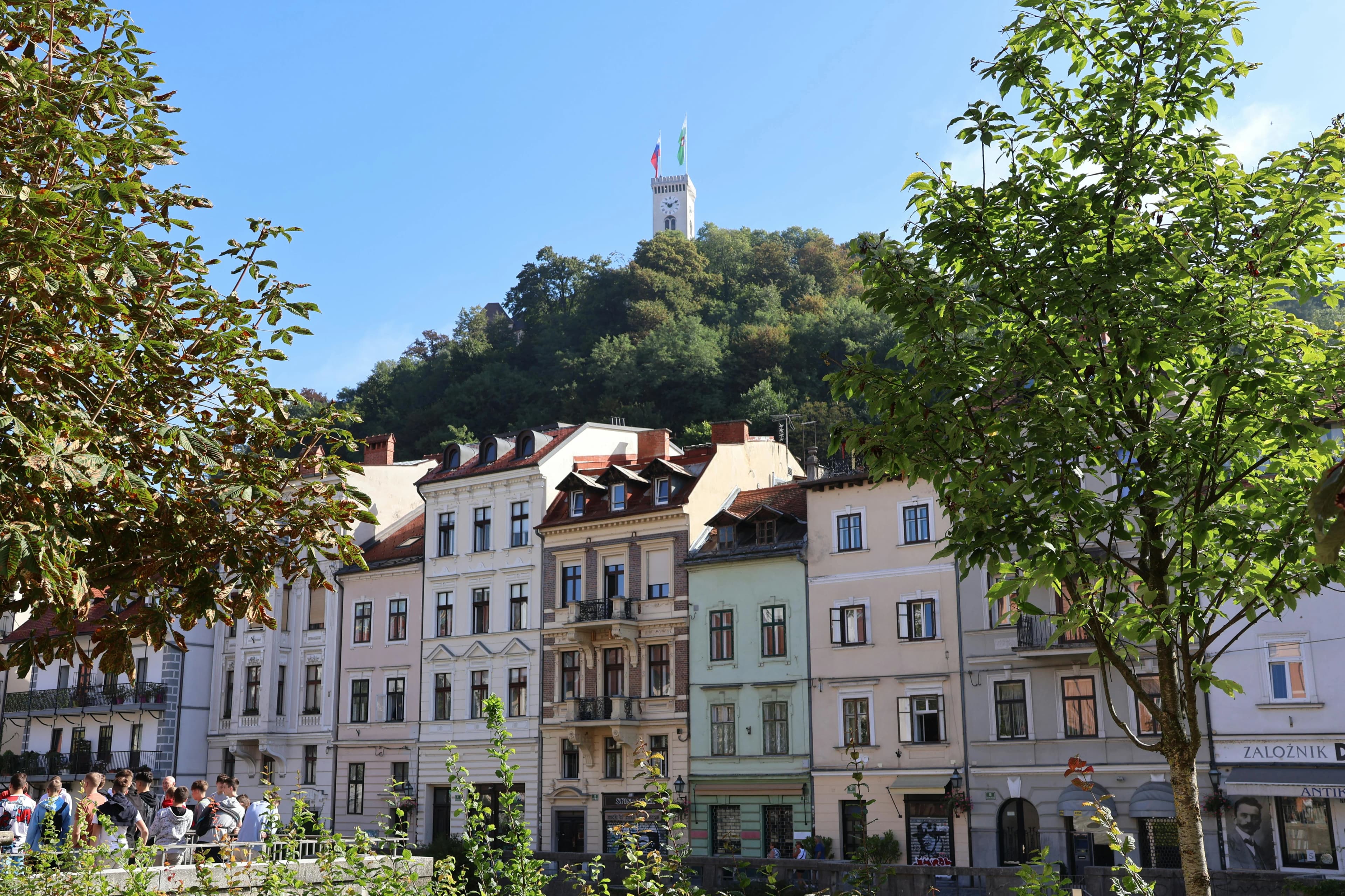 A row of traditional houses with colorful facades lines the Ljubljanica River, with the Ljubljana Castle tower rising from the hill in the background.