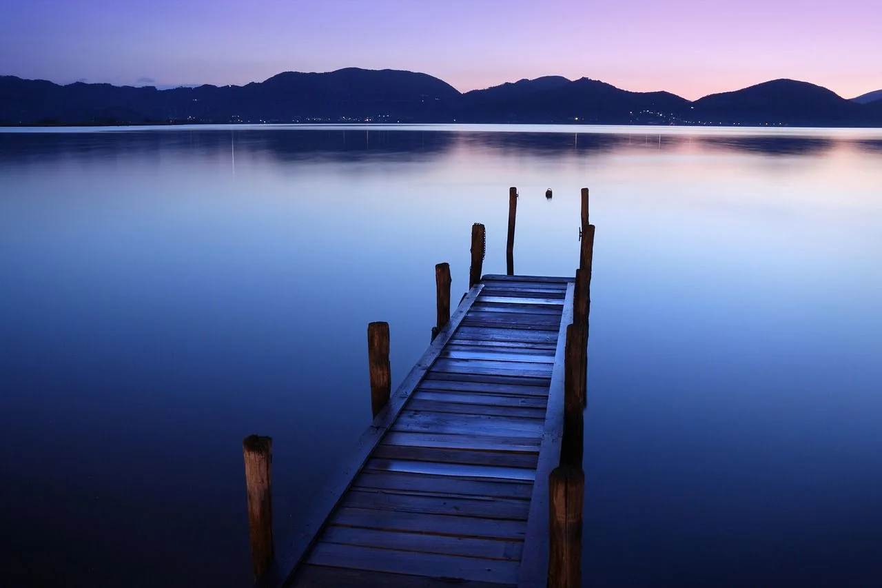 A quiet wooden pier stretches out into a calm lake at sunset, with the mountains silhouetted in the distance.