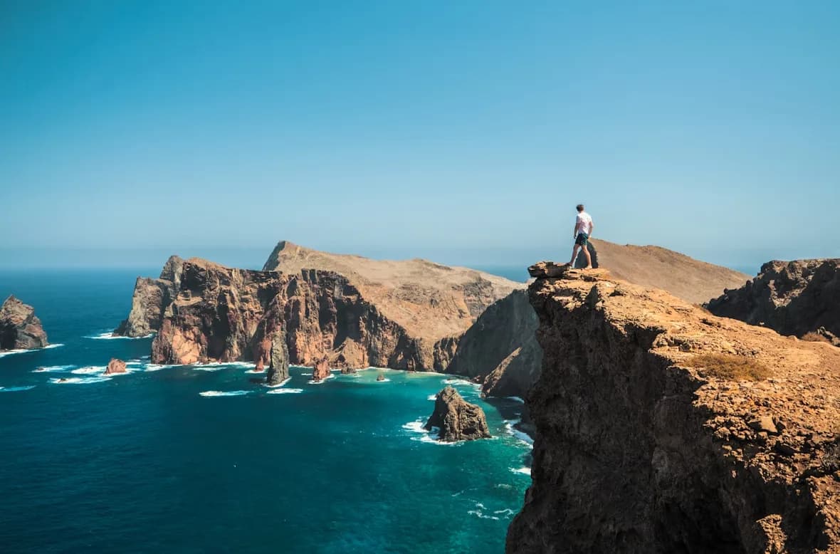 A person stands on a rocky cliff overlooking the ocean, with the dramatic, craggy coastline of Madeira in the background.
