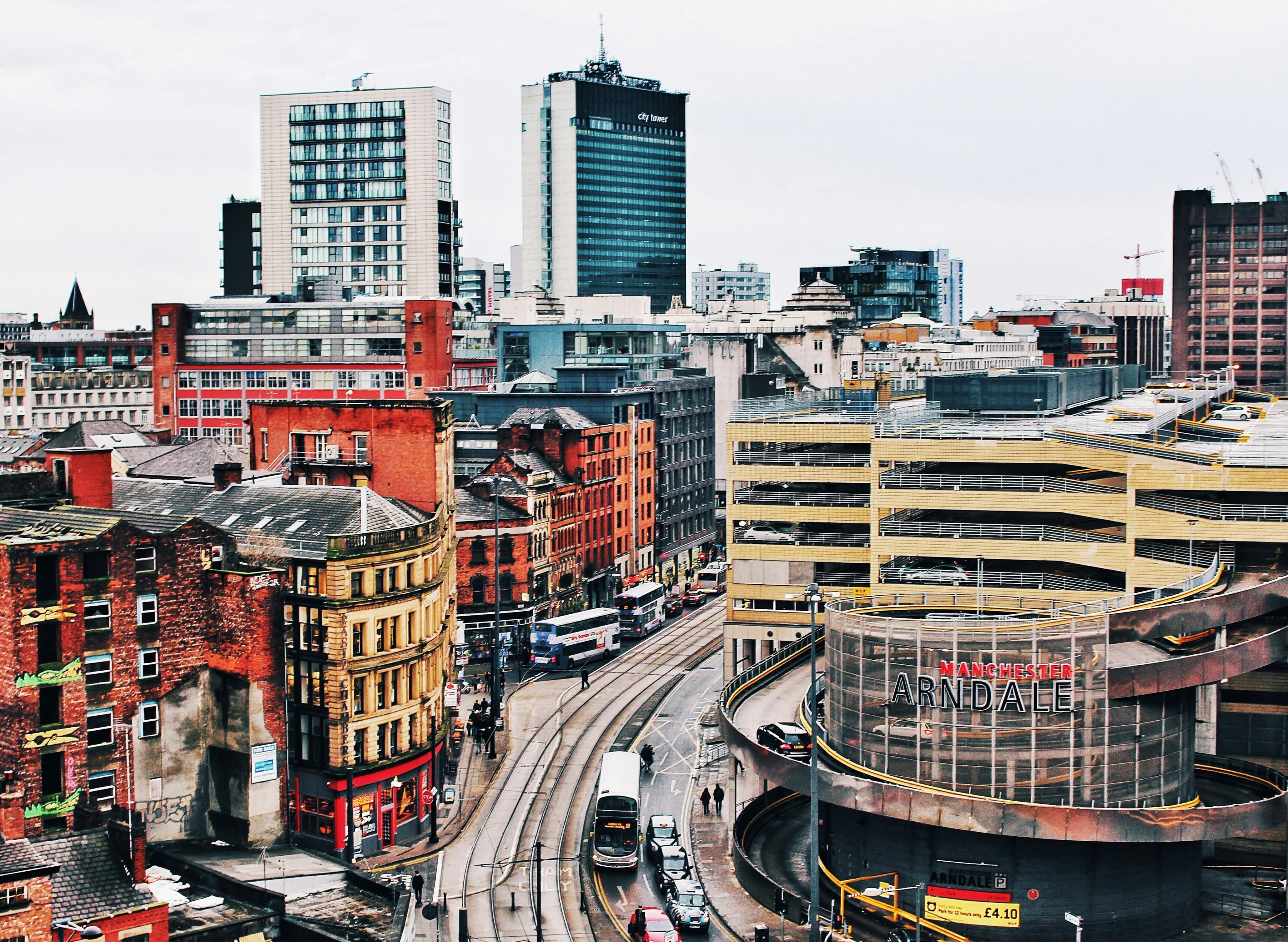 An aerial view shows the dense mix of historic and modern architecture in Manchester, with a bus and tram traveling along a city street.