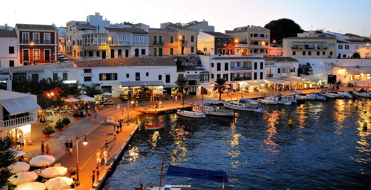 The picturesque harbor of Ciutadella is filled with boats, with the traditional white buildings of the city beautifully illuminated at dusk.