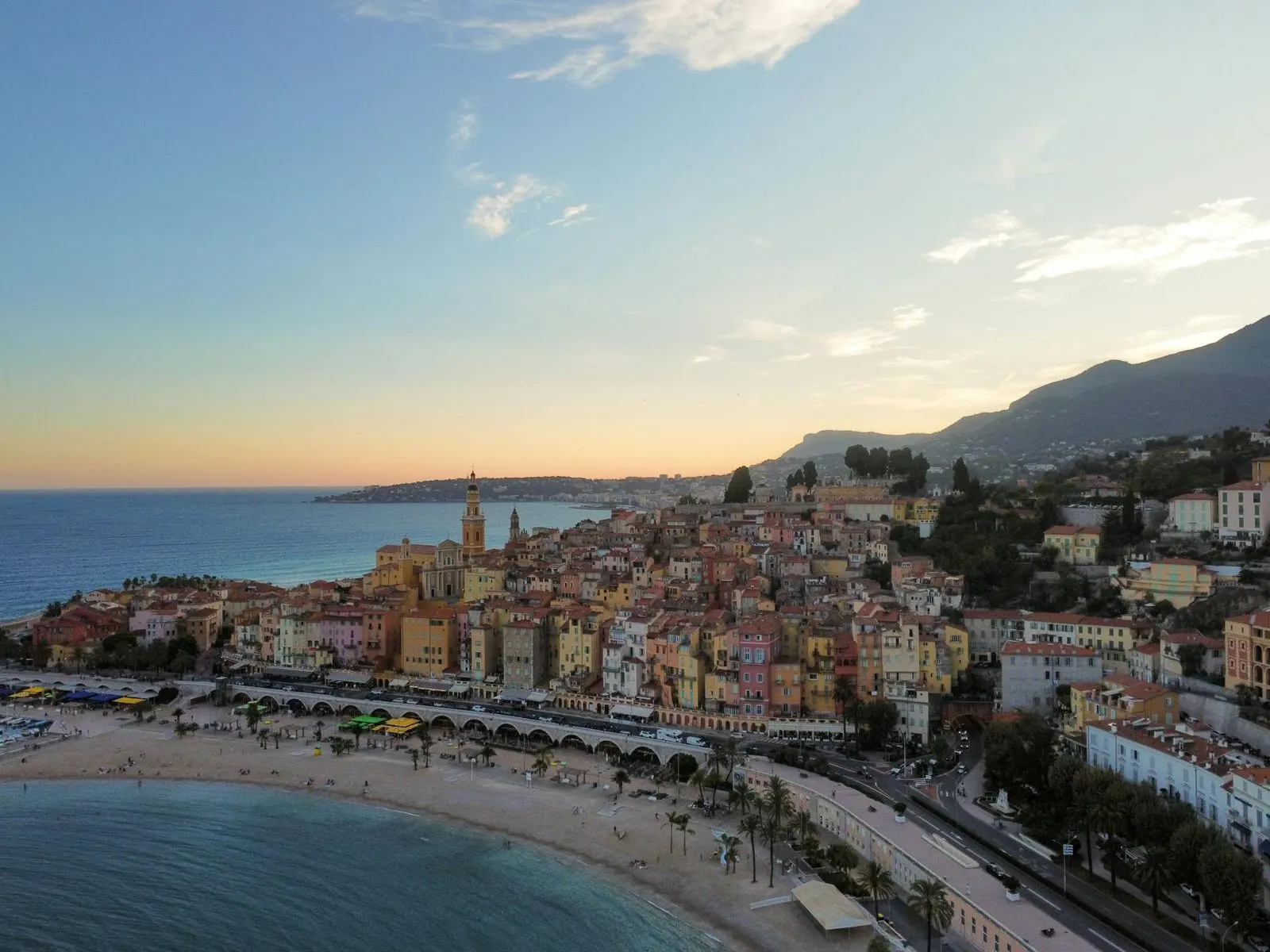 An aerial view captures the picturesque town of Menton, with its colorful buildings lining a sandy beach and a wide promenade.