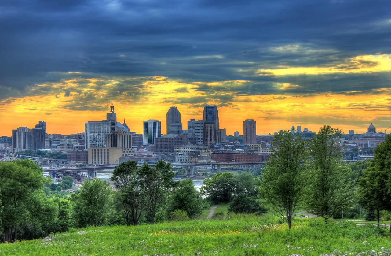 The Minneapolis skyline is silhouetted against a vibrant orange and yellow sunset, with a green park in the foreground.