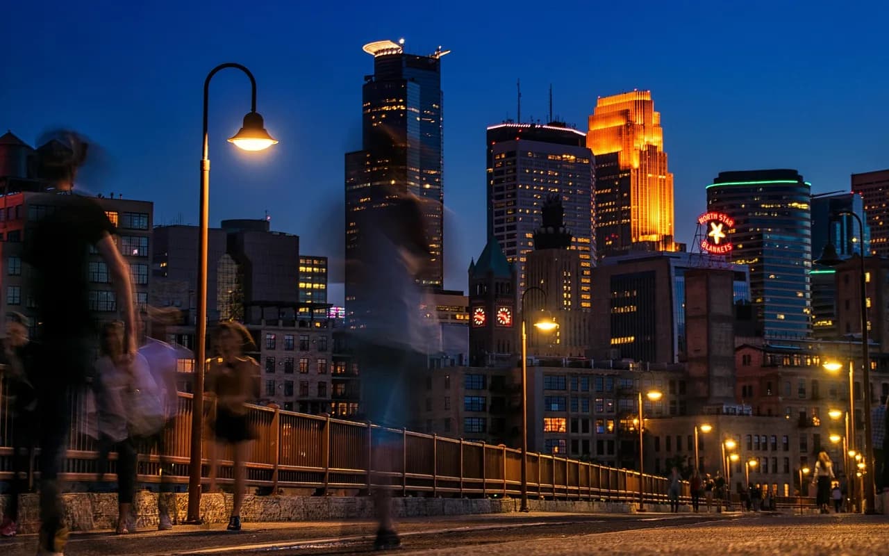 The Minneapolis skyline is illuminated at night, with the city lights and a vibrant neon sign glowing against a dark blue sky.