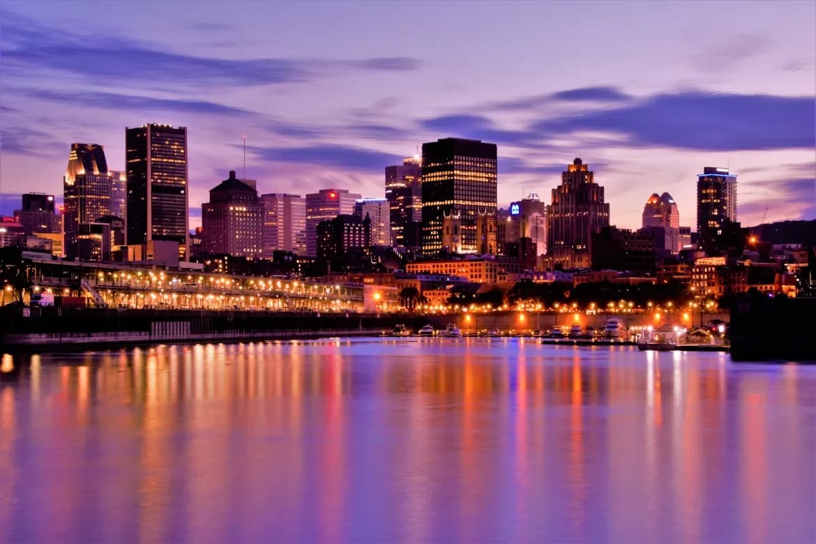 The Montreal skyline is beautifully illuminated at dusk, with the lights of the buildings reflected in the calm waters of the Saint Lawrence River.