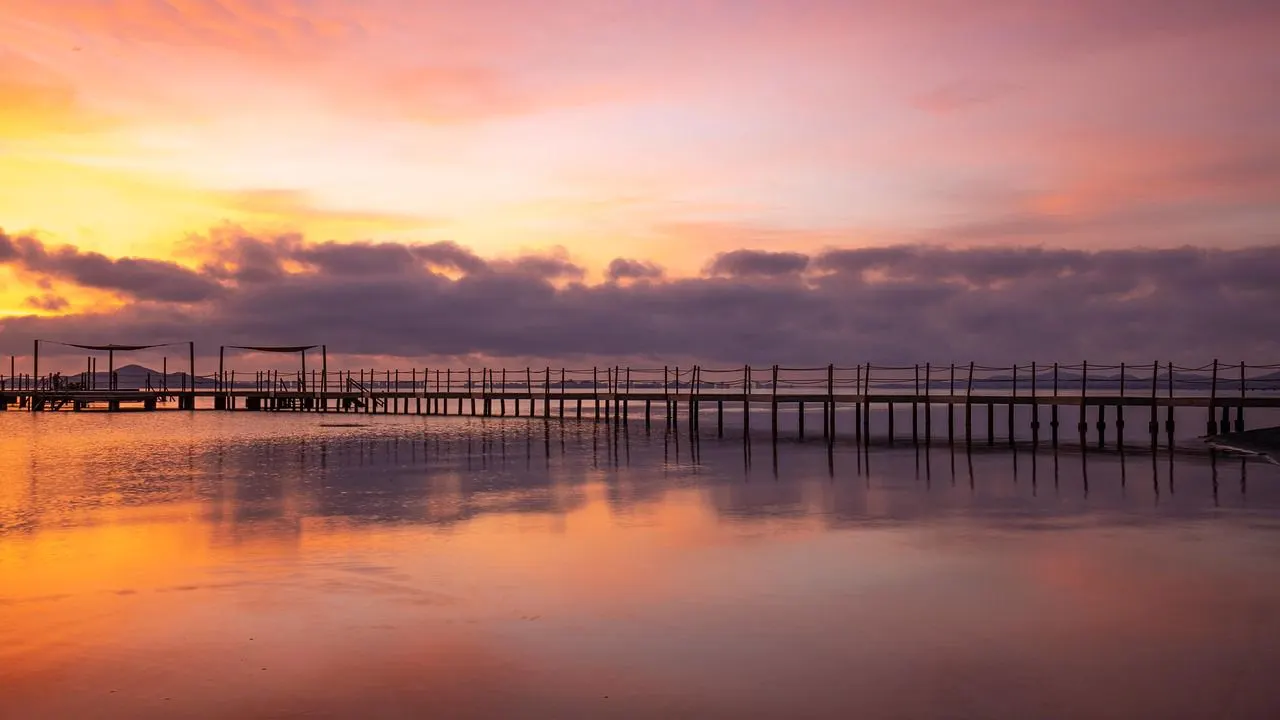 A long, wooden pier stretches over a calm lake at sunset, with a golden and purple sky reflected on the water.
