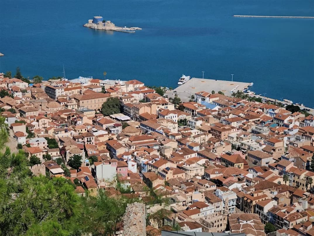 The dense rooftops of the historic town of Nafplion are viewed from above, with the Bourtzi Castle and the sea in the background.