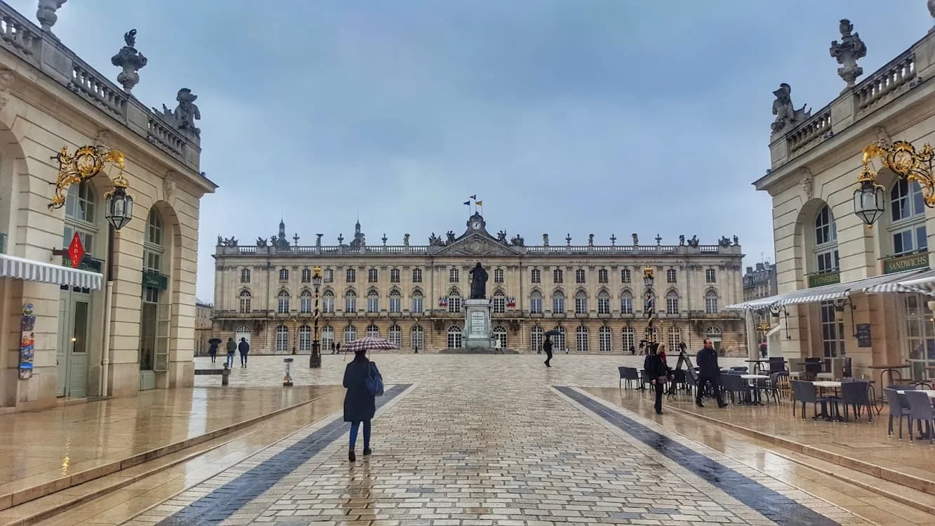 The magnificent Place Stanislas is viewed on a cloudy, rainy day, with the historic buildings and a statue in the square.