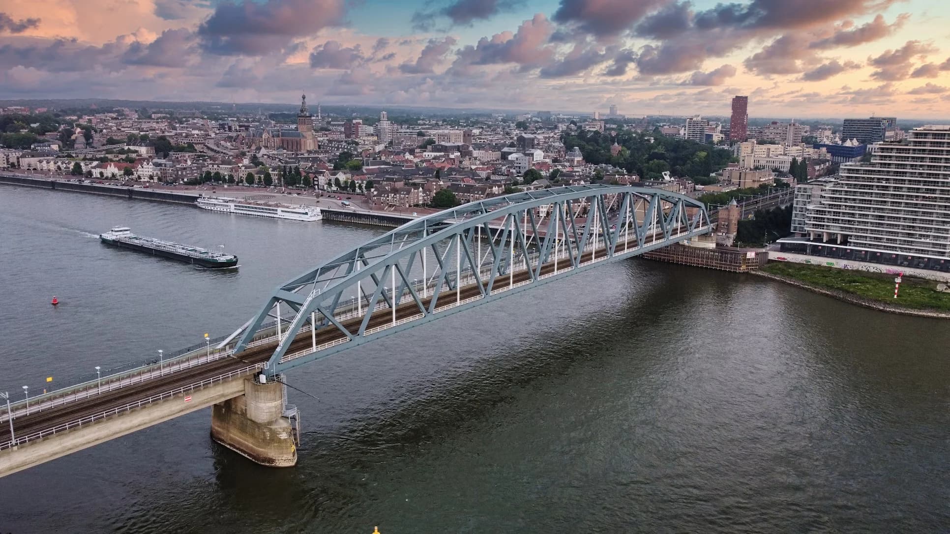 A panoramic view of the Nijmegen city skyline shows a mix of historic buildings and a modern bridge spanning the Waal River.