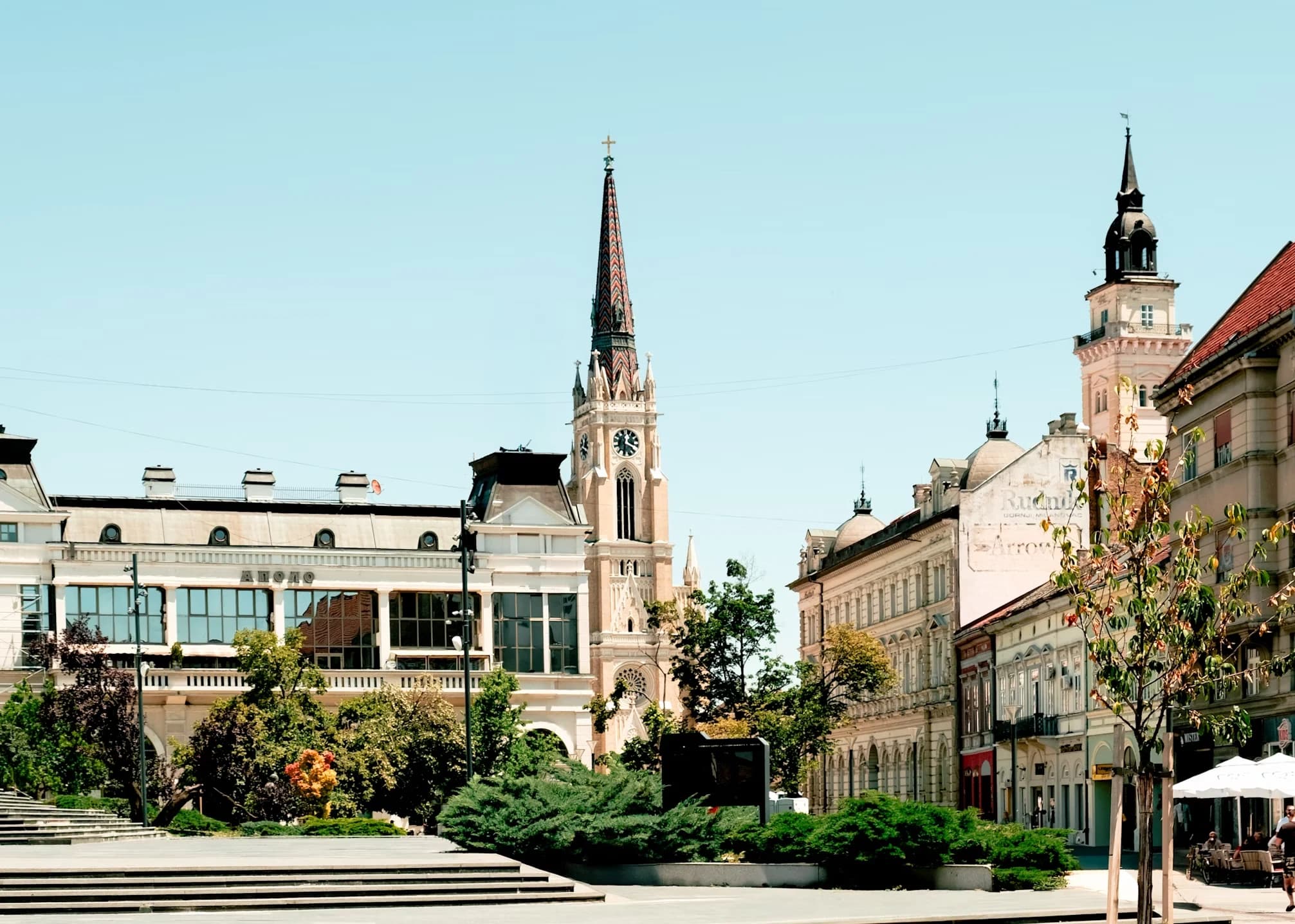 A large, historic church with a soaring steeple and a clock stands in the center of a city square.