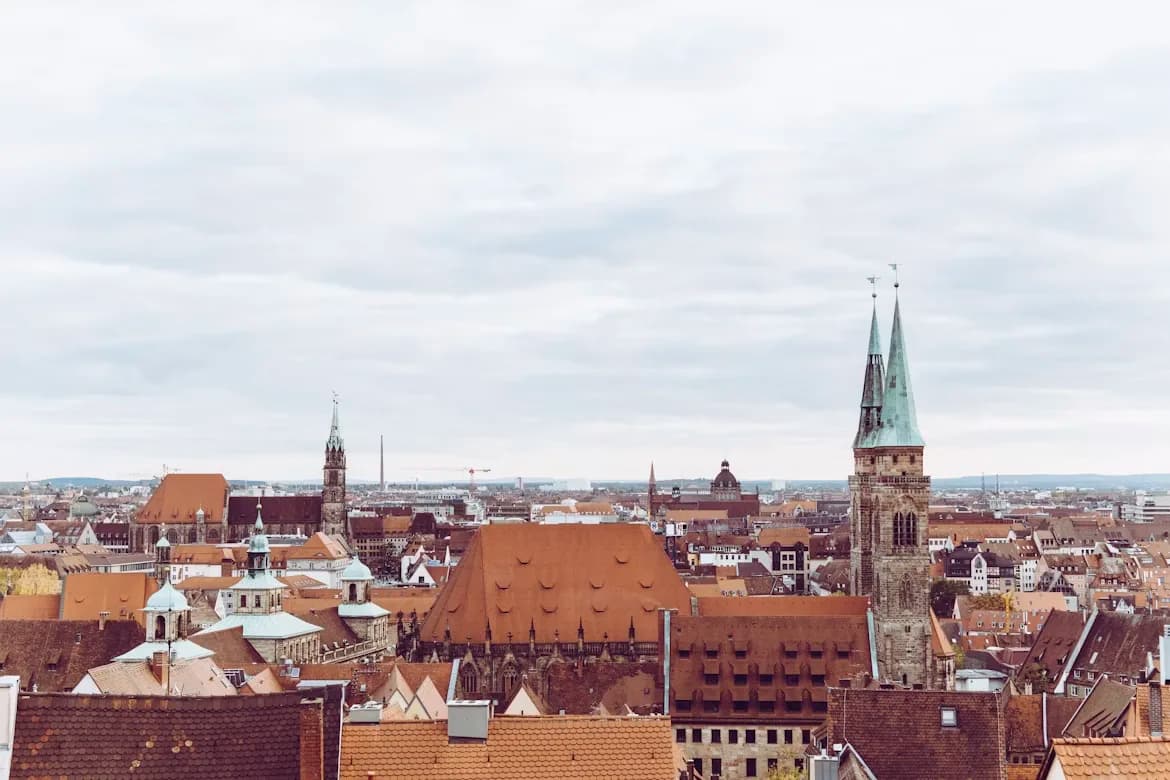 A panoramic view of Nuremberg's historic city center shows a sea of red rooftops and church spires under a cloudy sky.
