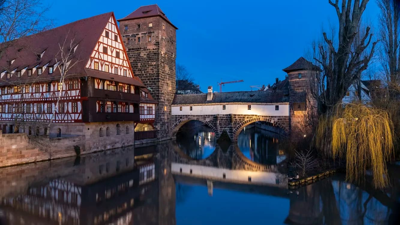 The historic half-timbered houses and a stone tower of Nuremberg are beautifully illuminated at night, with their reflections on the river.