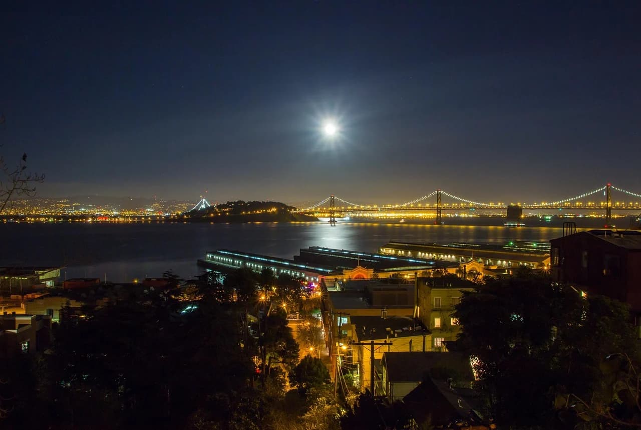 The San Francisco Bay and the illuminated Bay Bridge are a stunning sight at night, with the full moon shining brightly.