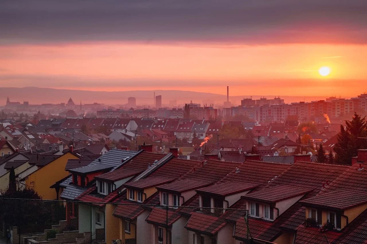 The sun sets over the rooftops of Olomouc, with the city's lights and distant hills glowing in the warm, orange light.