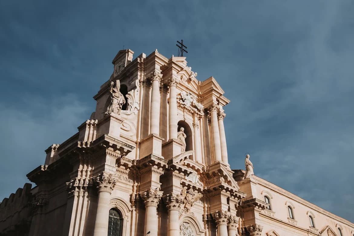 The ornate facade of the Duomo di Siracusa, a historic cathedral, is viewed from a low angle, with its detailed columns and statues against a cloudy sky.