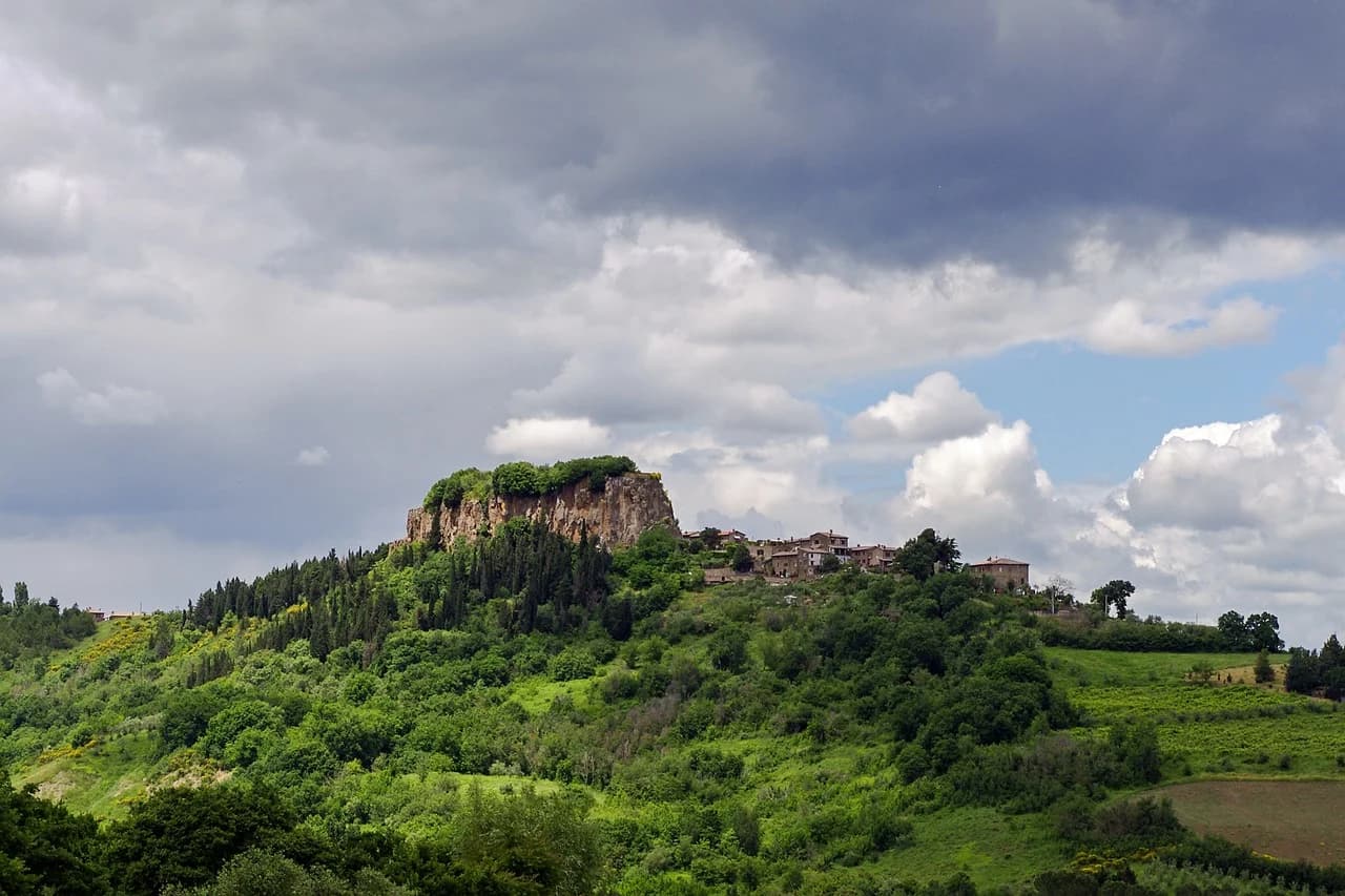 A historic village is perched on a hilltop, surrounded by lush green hills and a cloudy sky.