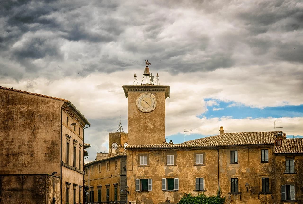 The medieval Torre del Moro, a clock tower with a large bell, stands in a city square, with historic buildings around it.