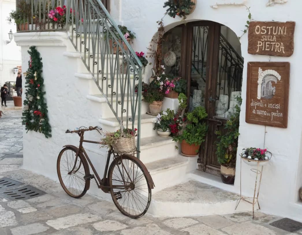 An old, rusty bicycle with a basket of flowers is propped against a whitewashed building with a staircase.