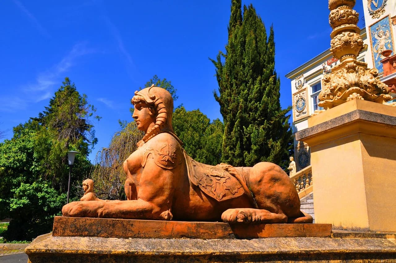 A large, ornate stone statue of a sphinx sits in a beautiful garden with a building and cypress trees in the background.