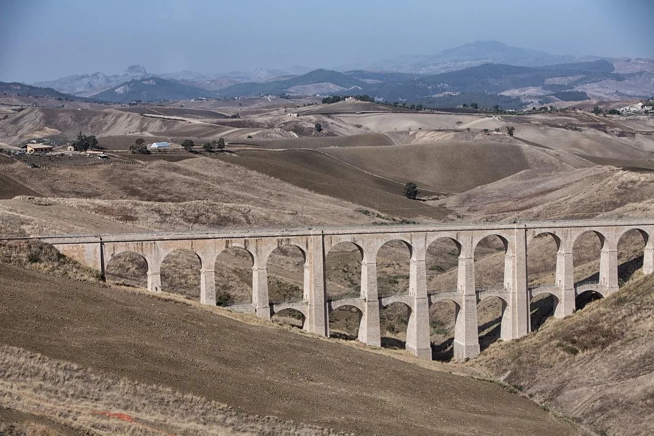 A historic stone aqueduct with numerous arches spans a dry valley, with rolling hills in the background.