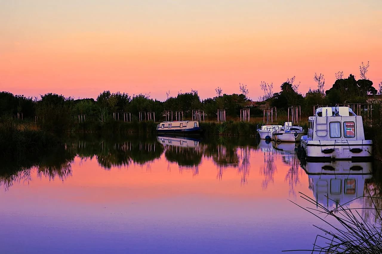 A small collection of boats is moored on a tranquil river at sunset, with the sky and water in beautiful shades of pink and purple.