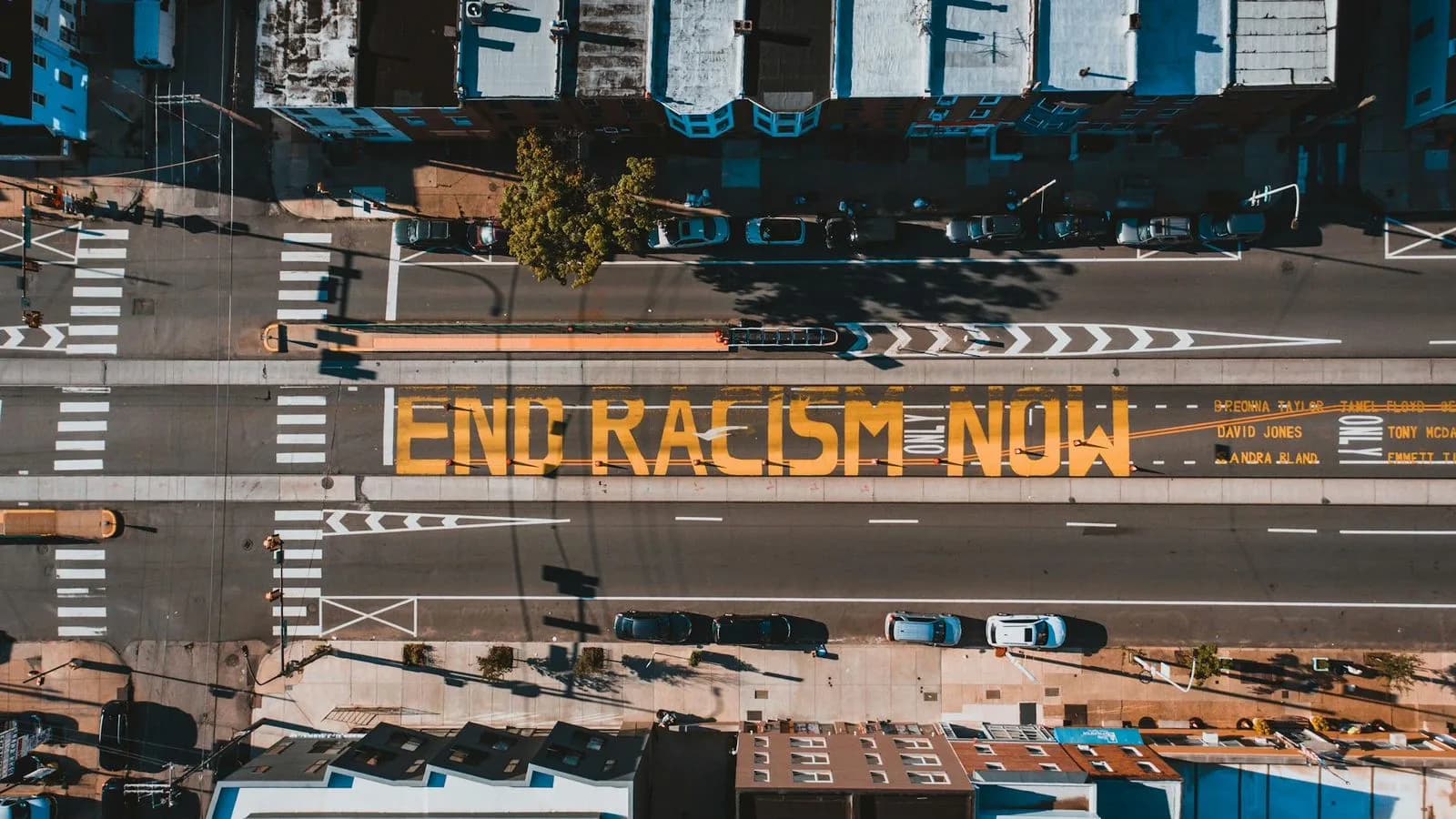 An aerial view captures a city street with a large yellow mural painted on the asphalt, with the words "End Racism Now."