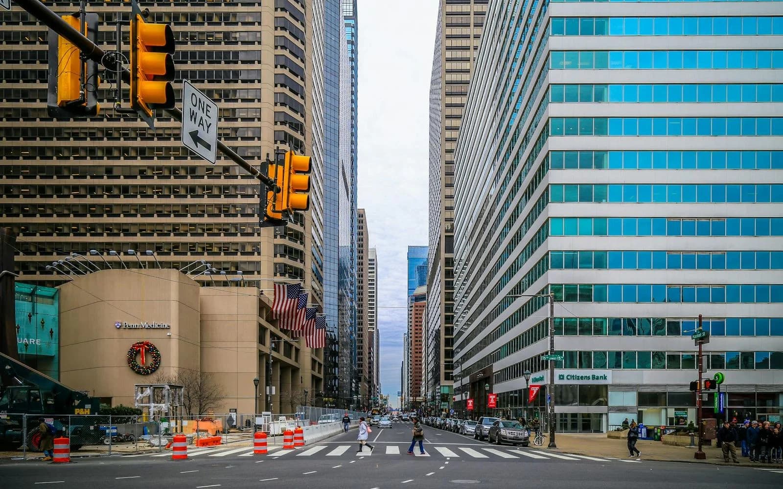 A busy city street is lined with modern skyscrapers, with a large traffic light and a Christmas wreath visible on a building.