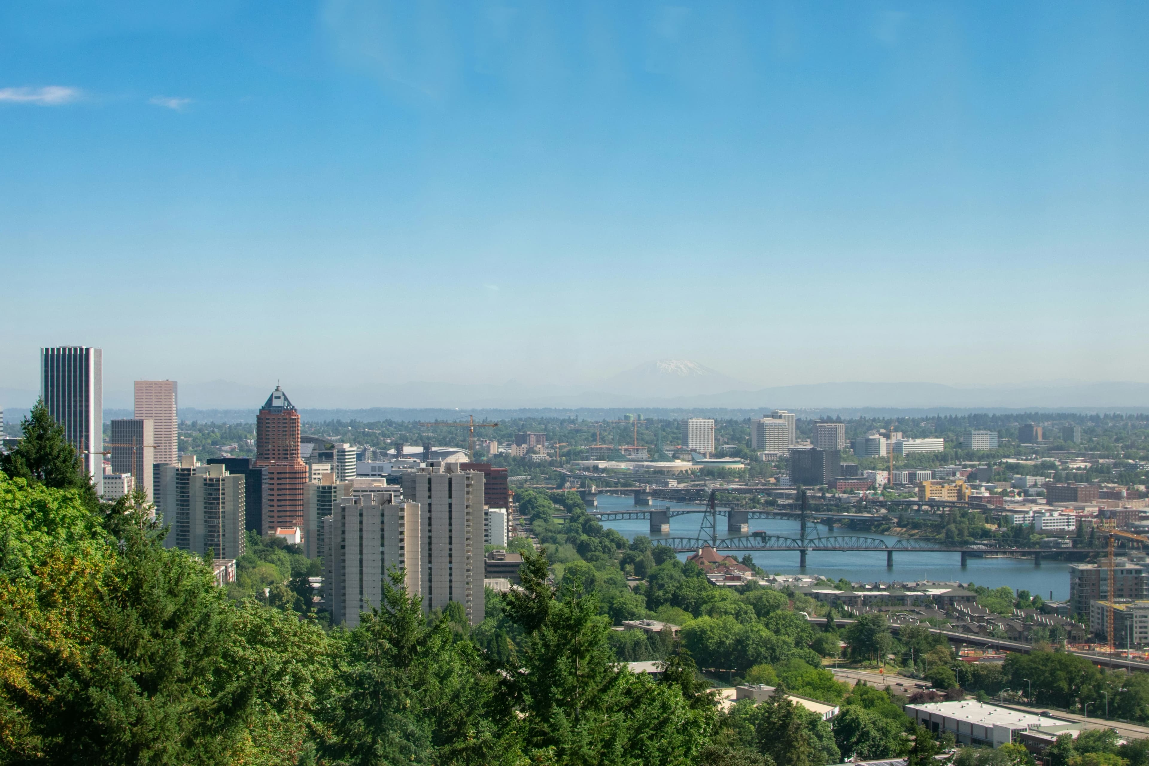 A panoramic view of the Portland skyline with the Willamette River and its bridges, with a distant view of Mount Hood.