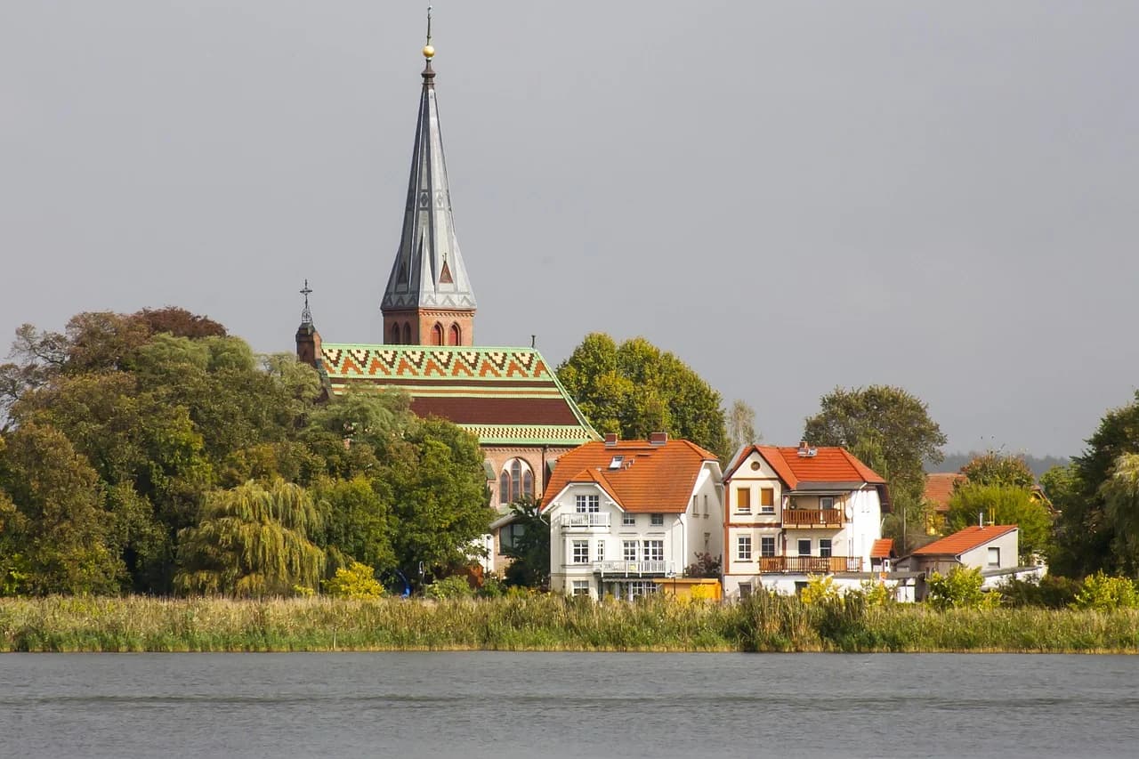 A historic church with a large steeple and traditional red rooftops stands by a calm lake.