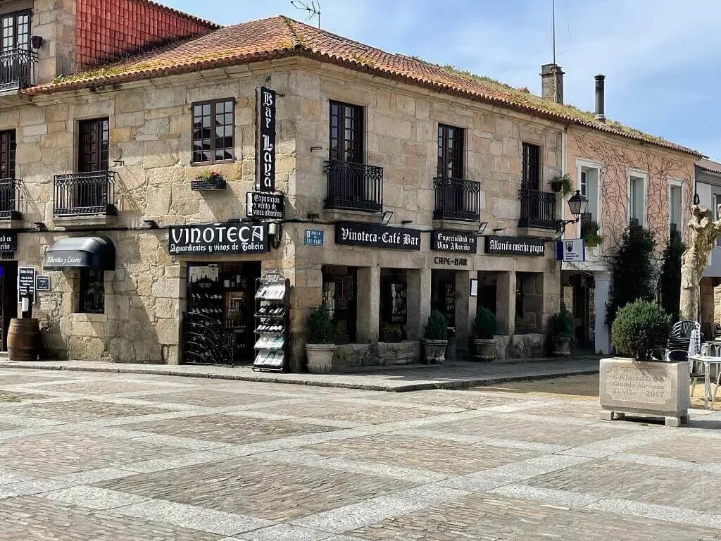 A stone building with a sign that says "Bar Loya" is a highlight of a city square, with people walking by and enjoying the day.