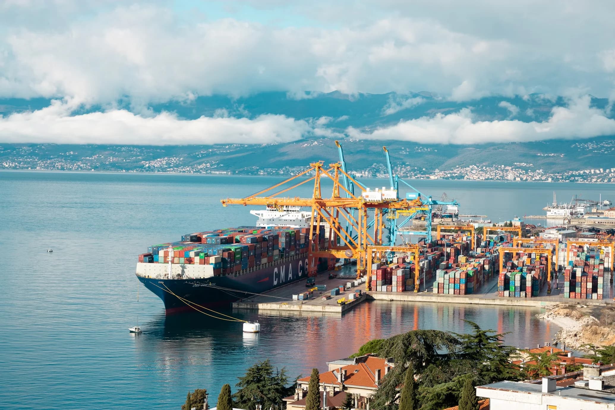 A massive cargo ship is docked in the busy port of Rijeka, with numerous cranes and shipping containers on the pier.