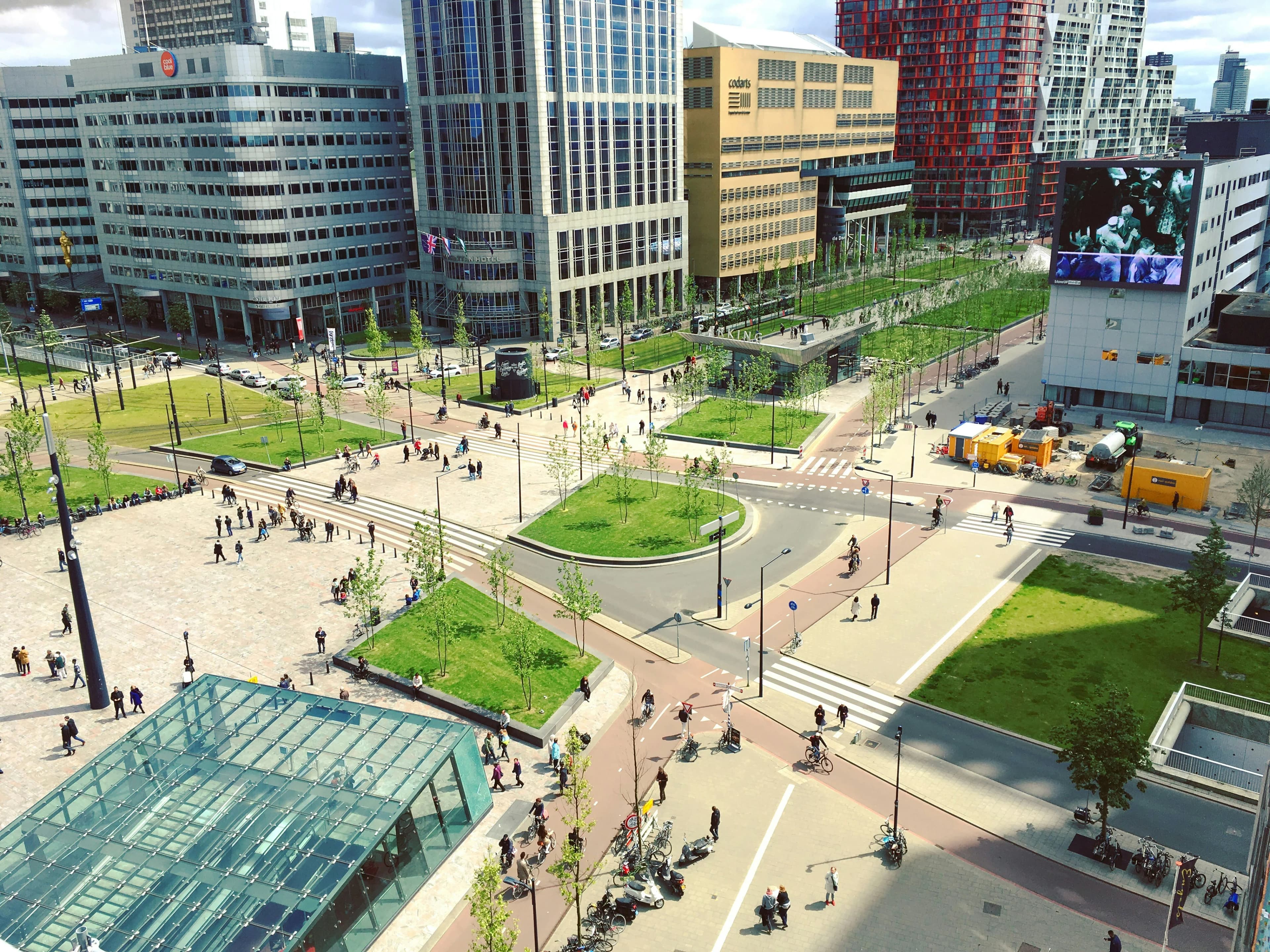 A large, modern square is bustling with people, bicycles, and various modern buildings in Rotterdam's city center.
