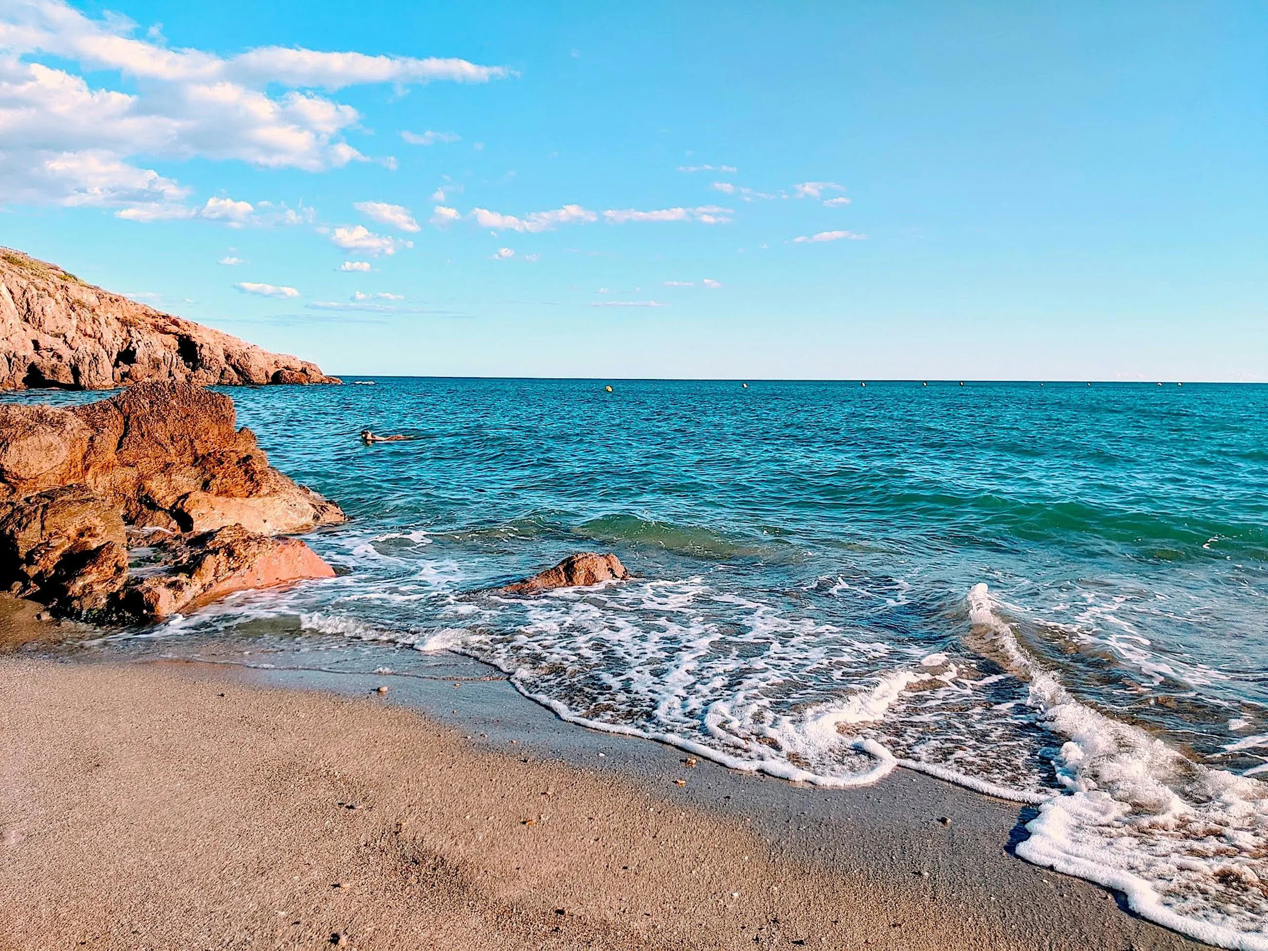 A small, sandy beach with a few rocky outcrops is bordered by a clear blue sea, with a few people swimming.