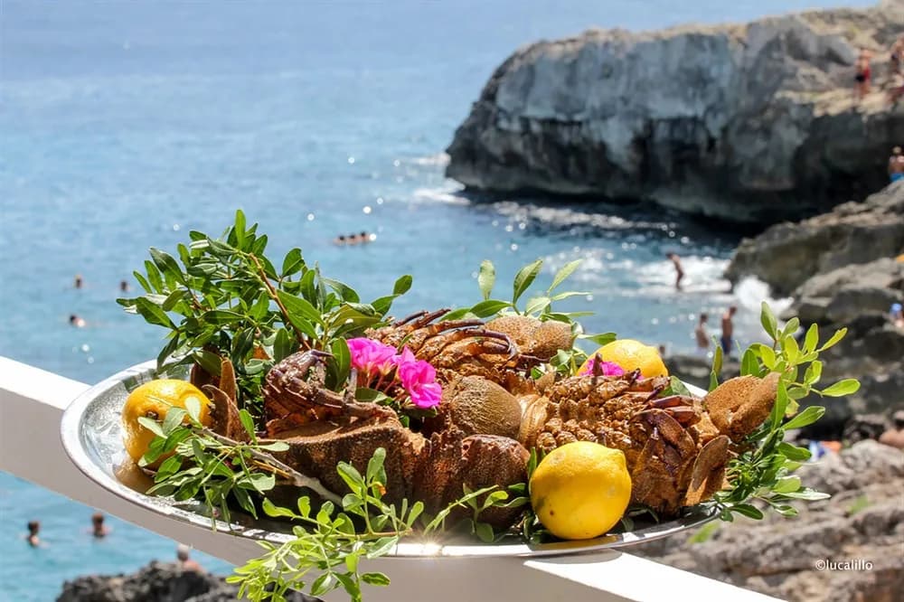 A large platter of freshly caught seafood is presented on a table, with a stunning view of the blue ocean and a rocky coastline in the background.