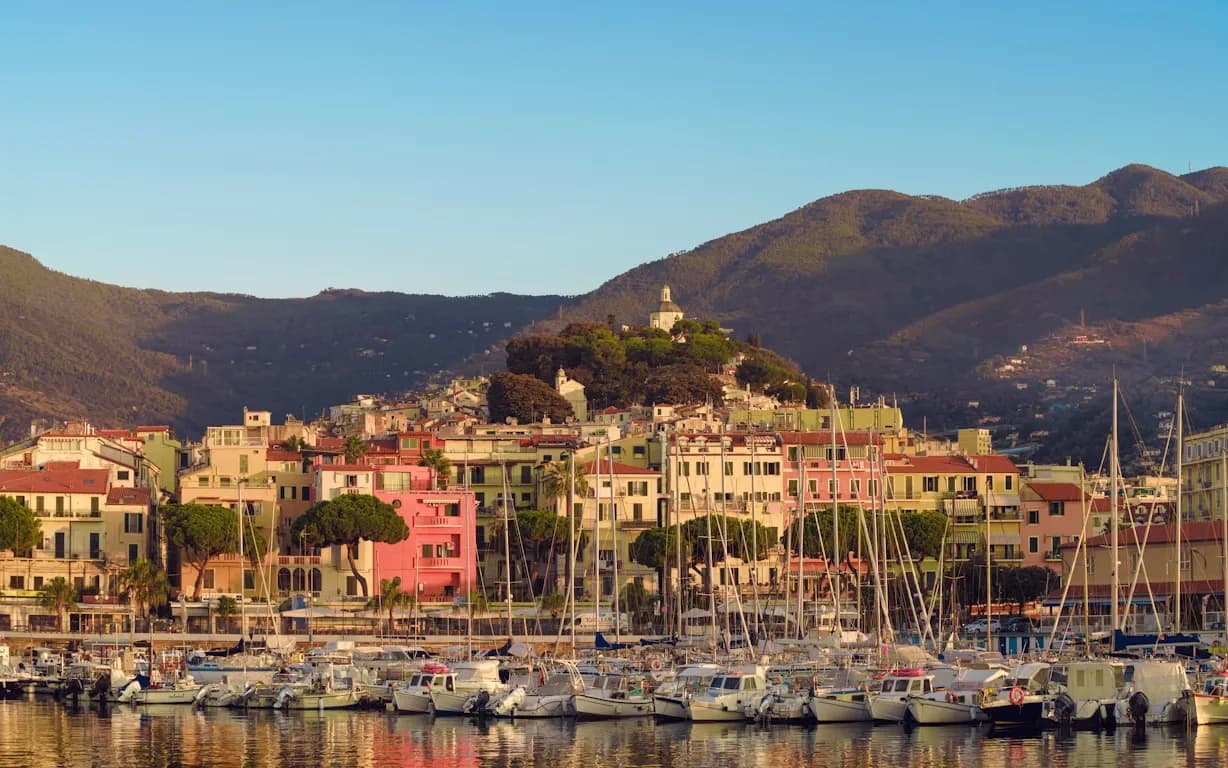 The colorful buildings of Sanremo's harbor are illuminated by the setting sun, with boats and yachts moored on the calm water.