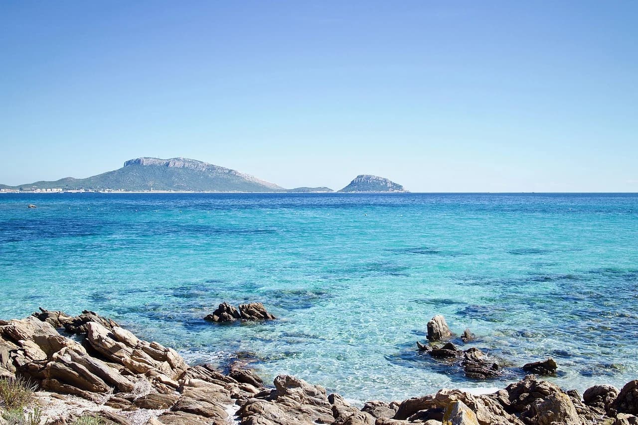 A wide, sandy beach with clear blue water is framed by a line of rocks, with mountains and a city in the distance.