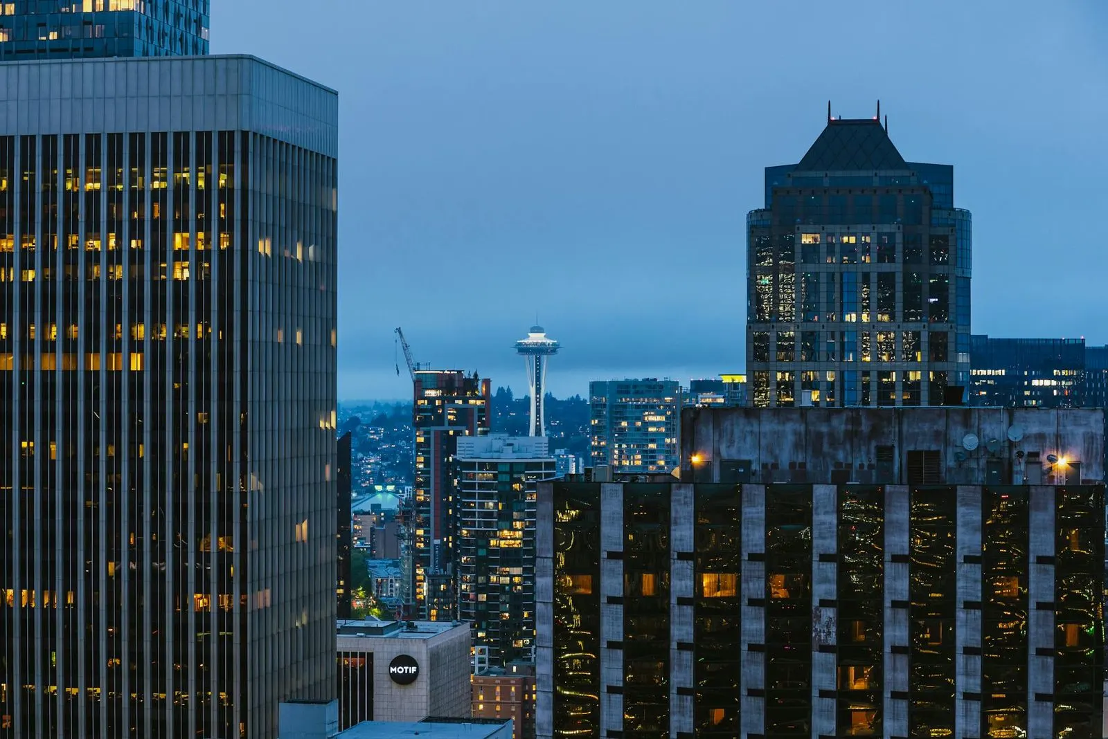 The Seattle skyline at dusk shows the Space Needle and other skyscrapers illuminated against a dark blue sky.
