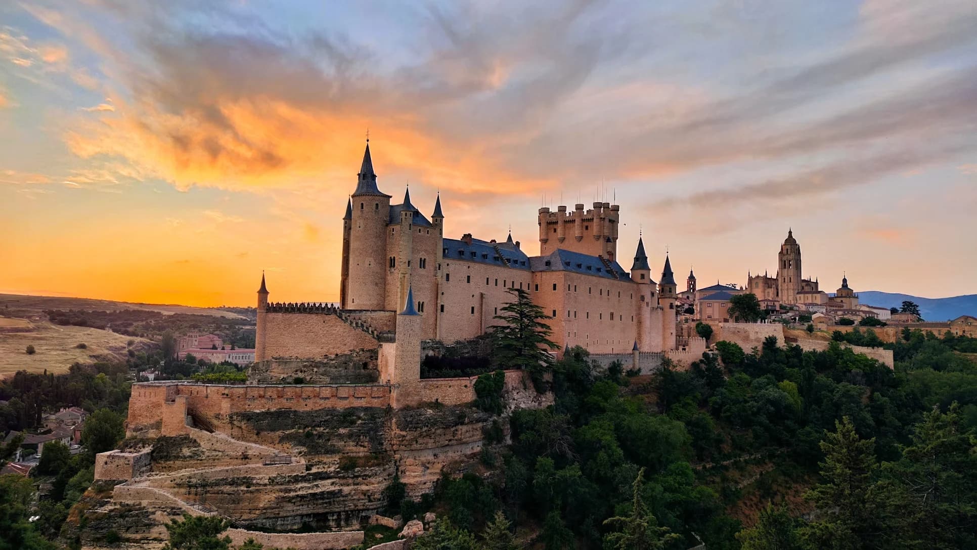 The iconic Alcázar of Segovia, a historic fortress with a pointed tower, is a stunning sight on a hill at sunset.