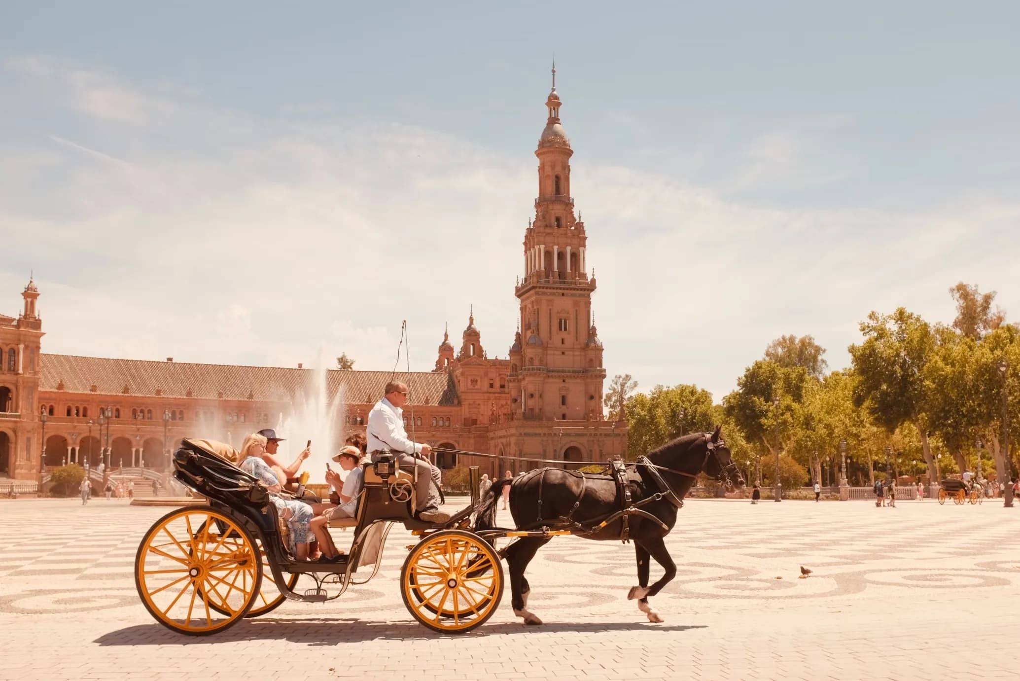 A horse-drawn carriage carries tourists through the Plaza de España, with the towering bell tower and fountains in the background.